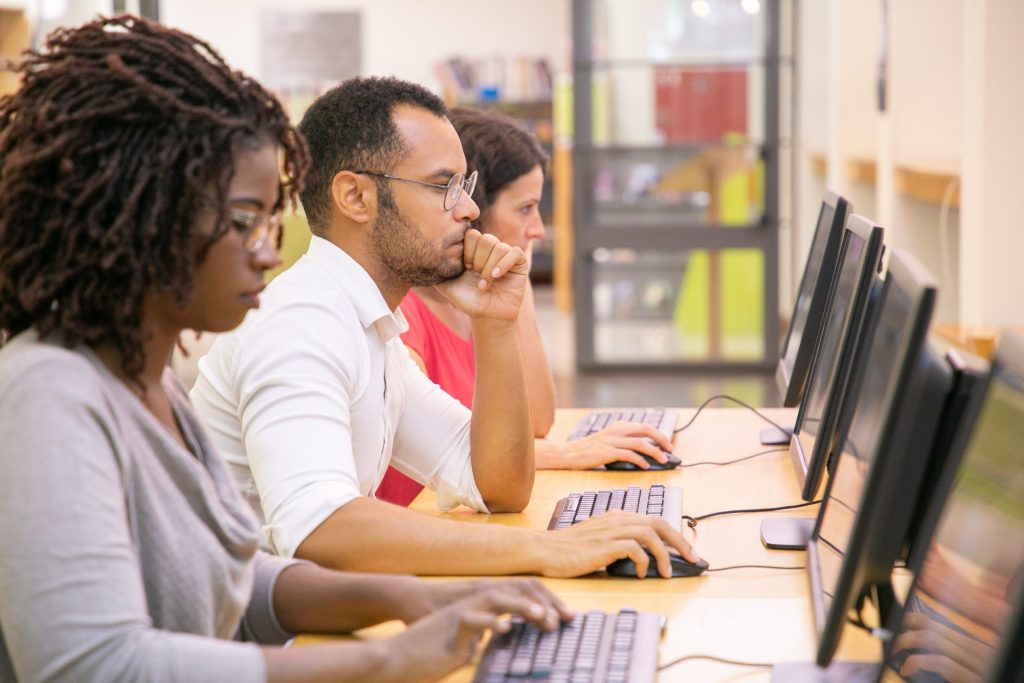 Grupo multirracial de alunos treinando na aula de informática. Linha de homens e mulheres sentados casualmente à mesa, usando desktops, digitando, olhando para o monitor. Conceito de centro de treinamento